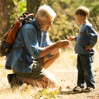 A grandfather with a backpack on kneels down to show a boy something on his hand. They are on a hike.