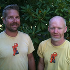 Two men in yellow uniform T-shirts pose in front of a leafy green background.