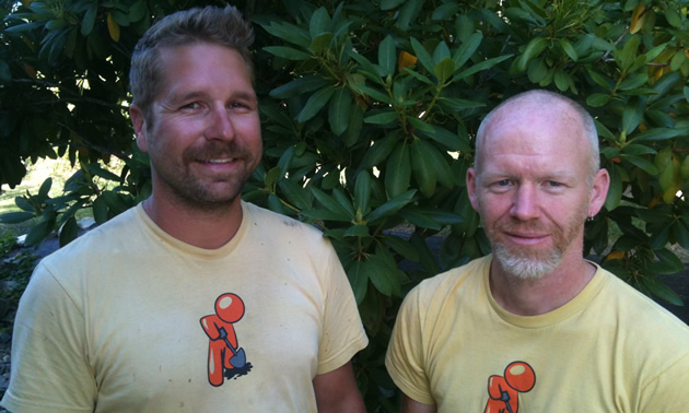 Two men in yellow uniform T-shirts pose in front of a leafy green background.