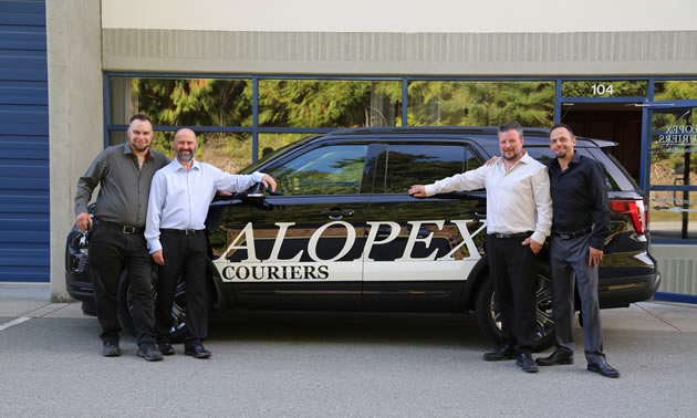 Four staff of Alopex Couriers stand in front of one of their fleet vehicles, a black car with the company's name in white letters on the side of the car.