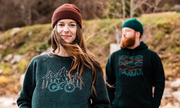 A woman standing wearing a dark green shirt and brown toque, man in background wearing dark green shirt and green toque. 