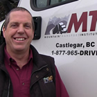 Broadly smiling man stands beside a white transport truck bearing the name Mountain Transport Institute, Castlegar, B.C.