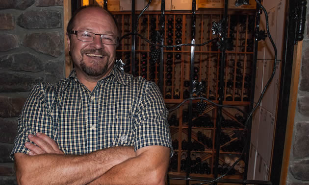 Bob Johnson, wearing a plaid shirt stands in front of an iron work gate leading to a wine cellar.