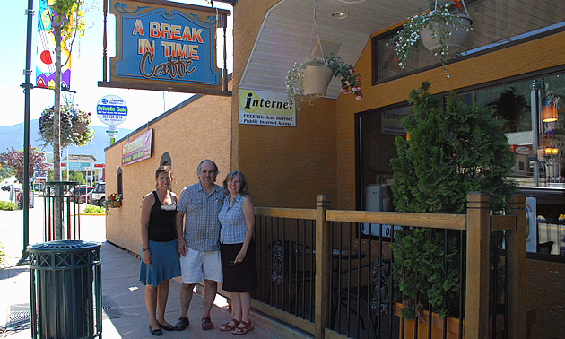 Three people stand beneath a wooden sign that Reads Break in Time Cafe. Behind them is a patio with greenery and an orange stucco building.