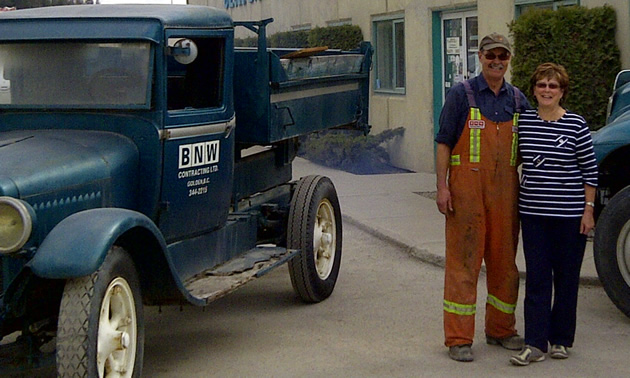 Smiling senior couple stand between an antique truck and a large haul-truck.