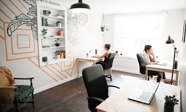 Sunny room with inset bookshelf and mural on wall, desks with two people sitting and working on laptops. 