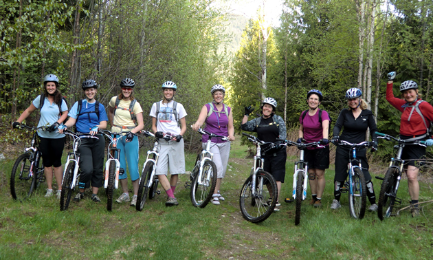 Women stand in a line on their mountain bikes against forest and trails.