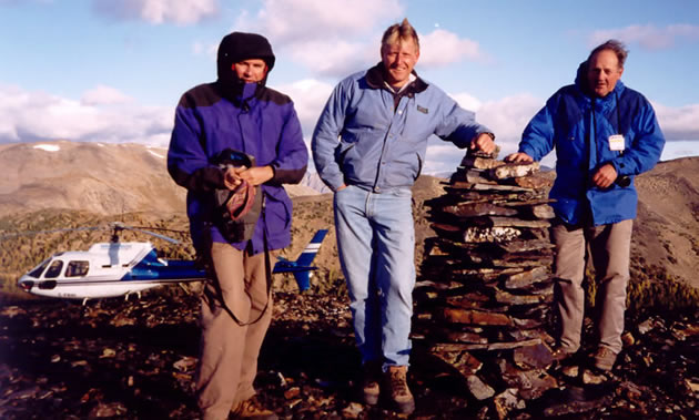 Three men stand beside a small tower of flat rocks; a helicopter sits in the background