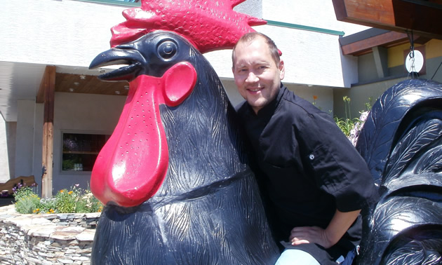 Smiling man sits astride a statue of a large black rooster