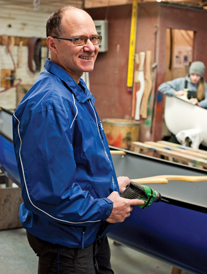 A smiling balding man stands with a drill ready to work on a canoe