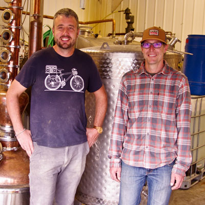 Wade Jarvis (L) and Trevor Semchuck standing in front of distillery equipment. 