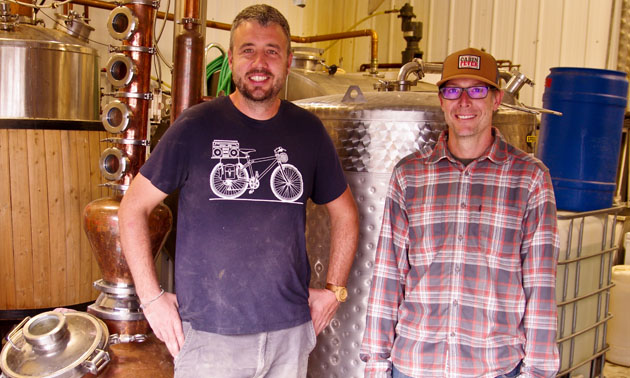 Wade Jarvis (L) and Trevor Semchuck standing in front of distillery equipment. 