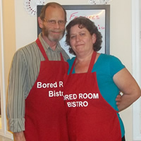 A couple stand with arms around each other. They both wear red aprons that read 