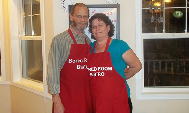 A couple stand with arms around each other. They both wear red aprons that read 