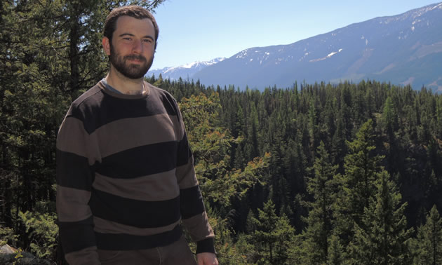 Brendan Murray-Nellis stands with evergreen forest, mountains and sky behind him