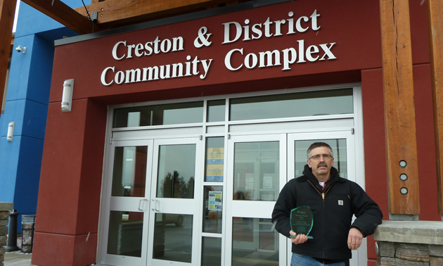 Man holding a clear acrylic trophy stands in front of the Creston & District Community Complex