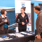 A man in a grey shirt talks to a recruiter who wears his navy fire gear and a ball cap. Behind them is an orange display and photos promoting Strategic Fire Control.