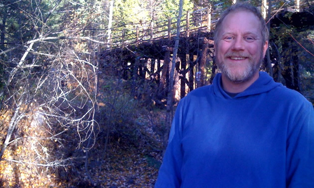 Smiling man stands in a wooded area in front of an elevated wooden footbridge
