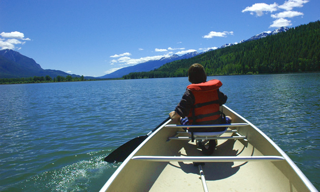 A boy sits in the front of a canoe. He is wearing a life jacket and paddles through still, blue waters.