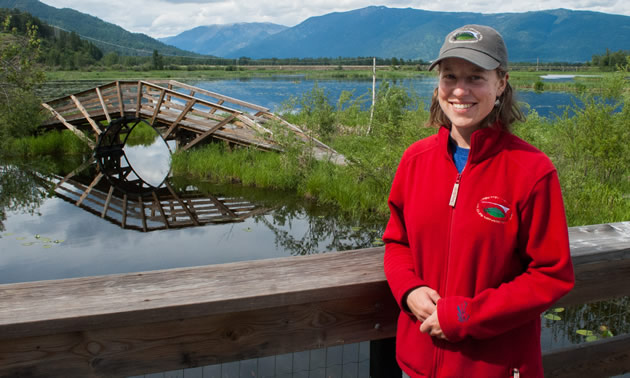 Andrea stands with a curved wooden bridge in the background. She wears a red fleece and a CVWMA cap.