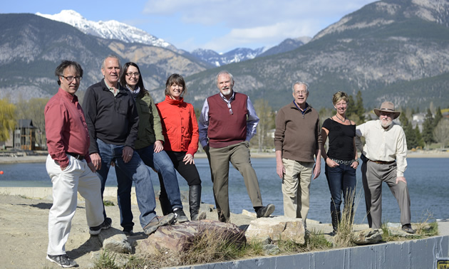 Eight adults stand on a rocky shore with water and mountains in the background