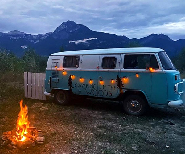 A blue and white VW van with lights strung across it, parked in front of a campfire. 