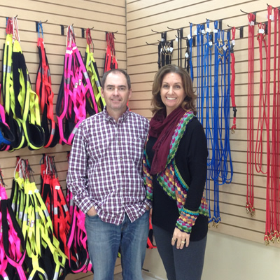 Rob and Shelley Ramsay, owners of Canadog in Kimberley, B.C., stand in front of a colourful selection of harnesses, collars and leashes.