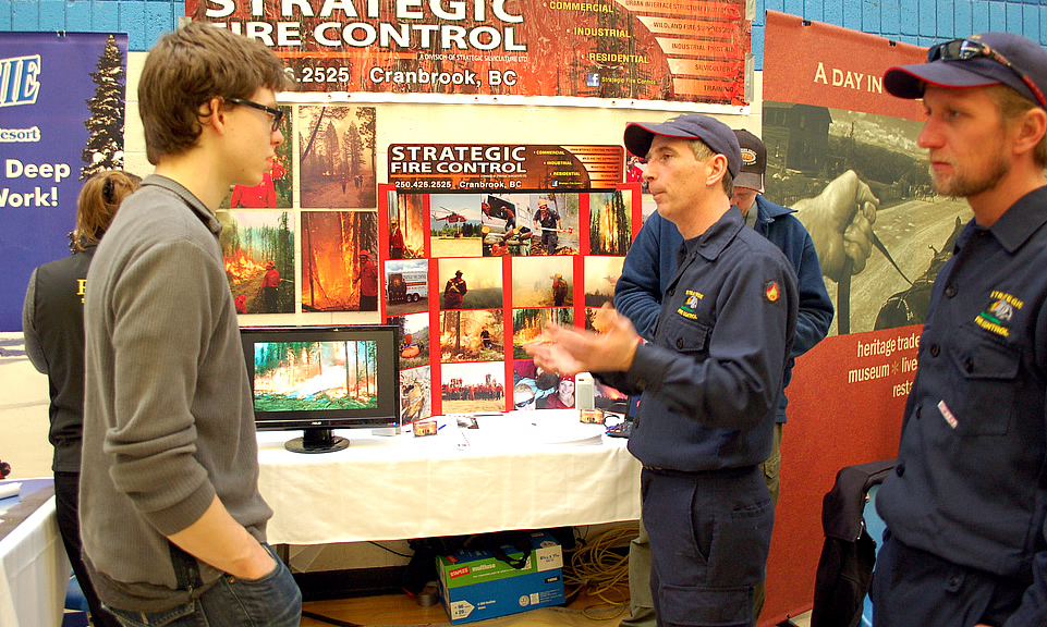 A man in a grey shirt talks to a recruiter who wears his navy fire gear and a ball cap. Behind them is an orange display and photos promoting Strategic Fire Control.