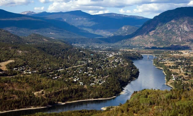 A view from a lookout looks over a river surrounded by mountians with Castlegar on either side.