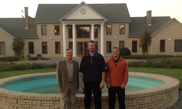 Three men stand in the foreground, with a circular pool and two-storey mansion behind them