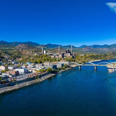 Aerial view of the City of Trail, showing wide river with a bridge across it, the city along the banks of the river and low mountains in the background. 