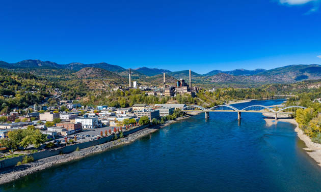 Aerial view of the City of Trail, showing wide river with a bridge across it, the city along the banks of the river and low mountains in the background. 