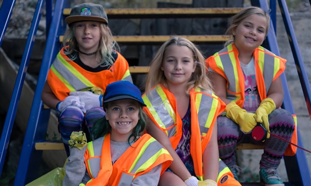 Four young girls in high-visibility vests sit on a wooden staircase with bags of trash