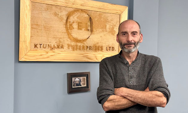 Corrie Walkley stands, arms folded in a boardroom in front of a wooden sign with the Ktunaxa Enterprises Ltd. logo. 