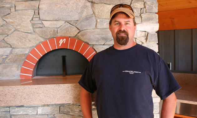 Man wearing a baseball cap and T-shirt stands beside a brick pizza oven