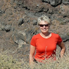 Smiling woman wearing red T-shirt and sunglasses stands waist-deep in grass on a rocky hillside above a body of water.
