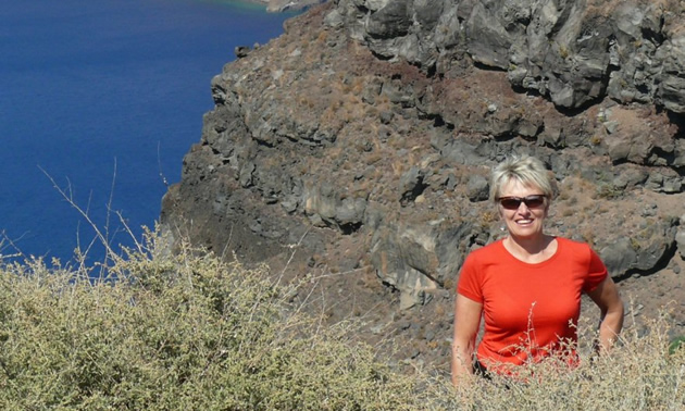 Smiling woman wearing red T-shirt and sunglasses stands waist-deep in grass on a rocky hillside above a body of water.