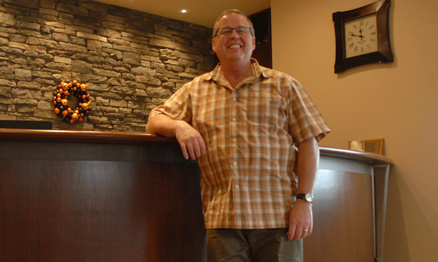 Dr. Nish is wearing a plaid shirt and glasses as he stands in front of a curved wooden counter front and rock work.