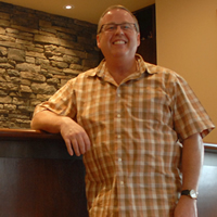 Dr. Nish is wearing a plaid shirt and glasses as he stands in front of a curved wooden counter front and rock work.