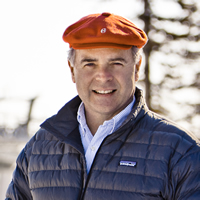 Man wearing a dark, quilted jacket and a bright red cap, stands on a snow-covered ski hill