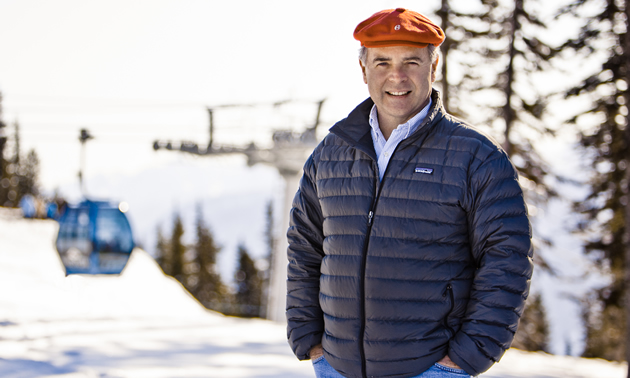 Man wearing a dark, quilted jacket and a bright red cap, stands on a snow-covered ski hill