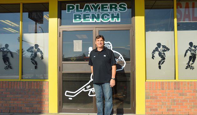 Man in a black sport shirt and jeans standing in front of a storefront, with the words Players Bench above the door