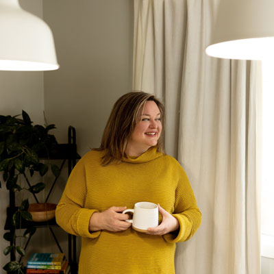 Dana Sproule standing by bright window looking outside, holding cup of coffee. She's wearing a mustard yellow sweater. 