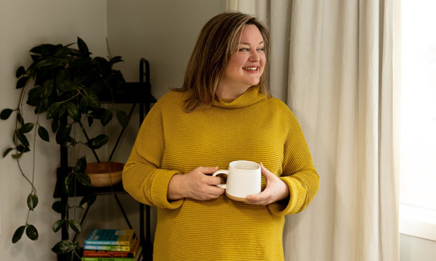 Dana Sproule standing by bright window looking outside, holding cup of coffee. She's wearing a mustard yellow sweater. 