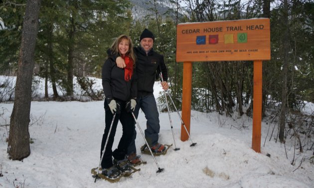 A woman and man wearing snowshoes stand beside a sign saying Cedar House Trail Head
