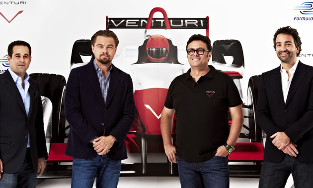 Leonardo Dicaprio and three other members of the Venturi team stand in front of a poster with logos and a life sized race car.