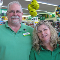Tall, grey-haired man with shorter blonde woman, both wearing green golf shirts and name-tags, with shelves of merchandise behind them
