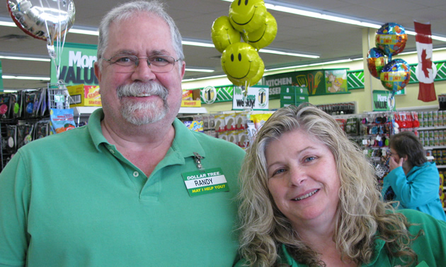 Tall, grey-haired man with shorter blonde woman, both wearing green golf shirts and name-tags, with shelves of merchandise behind them