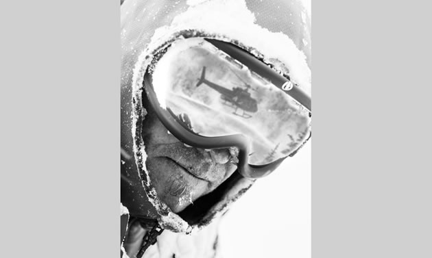 In this black and white photo a man's rugged face is framed by a snow covered hood. He wears snow goggles that reflect a helicopter.