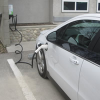 A white car is parked next to a blue and green charging unit that is plugged in.